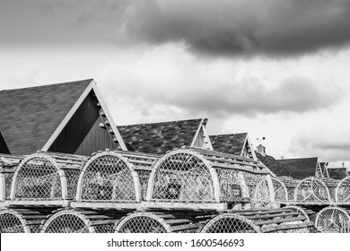 A Black And White Photo Of Stacks Of Weathered Wooden Lobster Traps In Front Of Wooden Sheds With White Trim In Bright Sunshine Under Dark Storm Clouds In A Fishing Village.