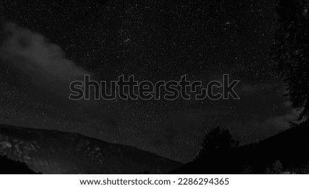 Black and white photo of a starry sky at night over a mountain and a forest with clouds in the trees in the Altai in Siberia.