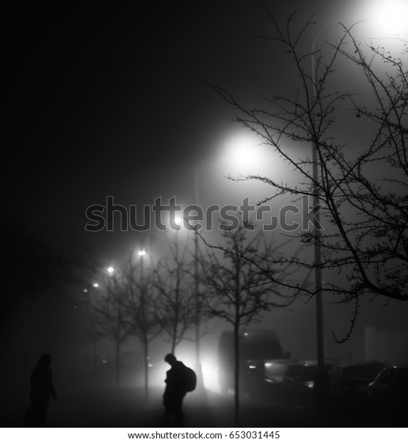 A black and white photo of a street at night with\
street lights shining behind the trees and silhouettes of two\
people on a foggy night