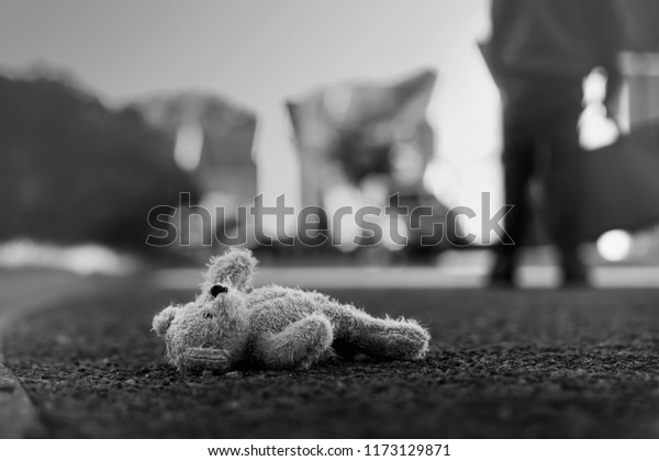 Black and white photo of Teddy bear laying  on\
the street with blurry background of school kid carrying school\
bag,Toy bear was left lying on the street,missing children or\
kidnap school kids\
concept