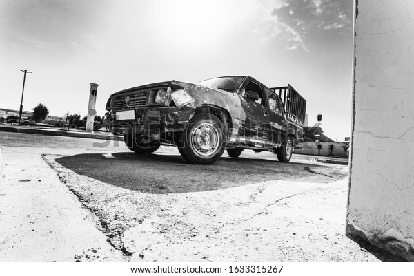 Black and\
white photo of a wrecked car with high contrast. A pickup truck in\
need of repair in a parking lot with a broken headlight. Scratched\
wing and car door after an\
accident