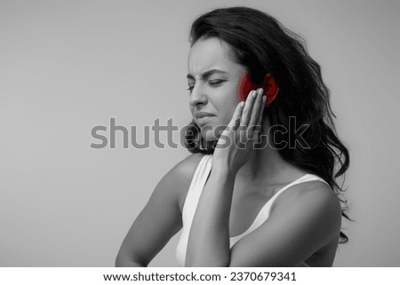 Black and white photo of young brunette eastern lady suffering from otitis, rubbing her inflamed ear highlighted with red over grey background, ear diseases concept, copy space