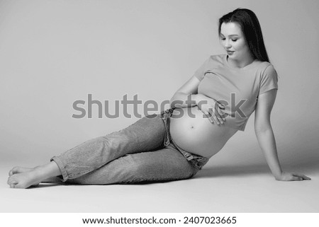 Black and white photo of young pretty pregnant woman in t-shirt and jeans sit on the floor on gray background. Female with belly exposed. 6th month of pregnancy.
