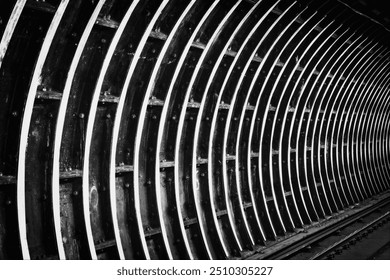 A black and white photograph of a curved tunnel wall featuring a series of parallel lines and metallic structures. The lighting creates a dramatic effect, emphasizing the depth of the tunnel. - Powered by Shutterstock