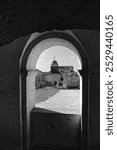 A black and white photograph showing the view through an arched window of Janowiec Castle, revealing its tower and ruins of the courtyard. Captures the medieval atmosphere of this historic Polish site