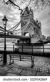 A black and white photograph of Tower Bridge in London with a park bench and cobblestone road in the forground