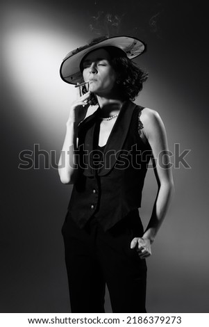 Black and white photography of stylish, elegant woman in classical black suit and stylish hat smoking, posing. Coco Chanel style. Monochrome effect. Concept of fashion, style, history