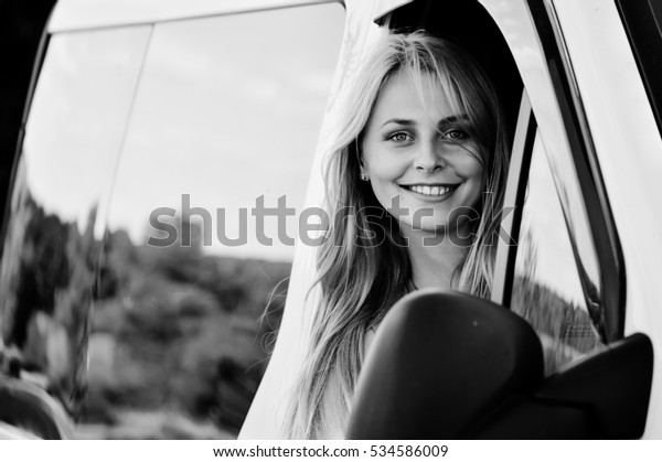 Black and white picture of beautiful woman\
driving car. joyful female looking from car window at camera on\
blurred countryside reflection\
background.