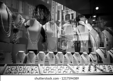 Black And White Picture Of A Display Of Pearl Necklaces And Rings In A Jewelry Shop Window.  Jewelry Store Glass Case Reflects City Buildings