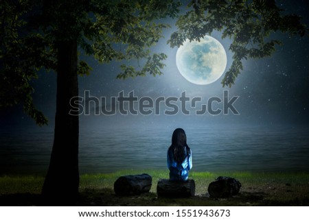 A black and white picture. The little girl sat and looked at the moon on a log by the sea on a full moon night beautifully.