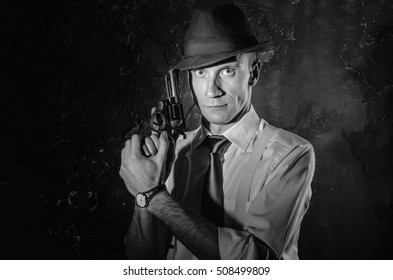 Black and white picture of a private detective with a gun in both hands. Agent in stylish hat. Man stays front to camera. He wears classic shirt with sleeves rolled to the elbow, suspenders, dark tie