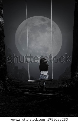A black and white picture of a young woman riding a swing looking at the full moon in the lonely night