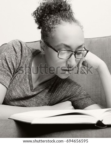Black and white portrait of beautiful african american student reading book wearing glasses laying on home sofa, serene thoughtful indoors. Black female college learning. Recreation hobbies lifestyle.