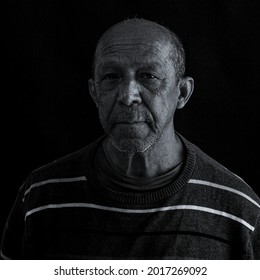Black And White Portrait Of An Elderly Man With A Sad And Tired Facial Expression Staring Straight Ahead On A Black Studio Background.