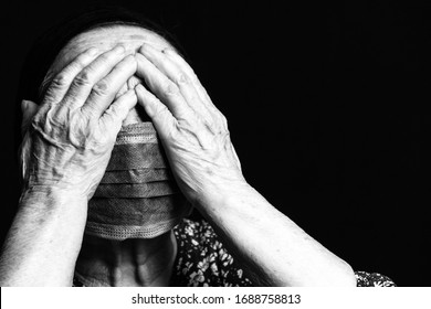 Black And White Portrait Of An Elderly Woman In Medical Mask Covering Her Eyes With Her Hands On A Black Background.  Old People In Risk Zone Because Of Virus Epidemy, COVID-2019 Pandemic