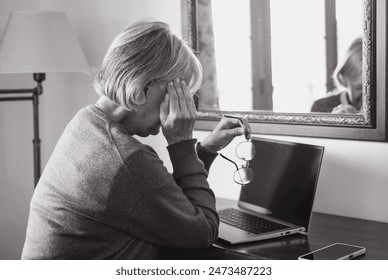 Black and white portrait of elderly woman using laptop at home while touching her temples for headache. Excessive work or study generates severe migraine - Powered by Shutterstock