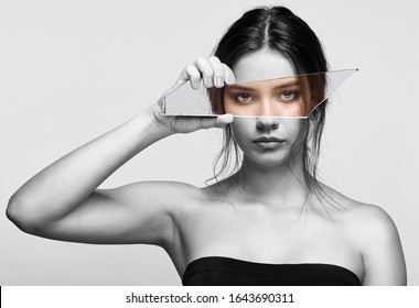 Black and white portrait of female with mirror shard in hand posing on gray background. Girl covers her face with a shard of the mirror. Eyes reflection in mirror splinter