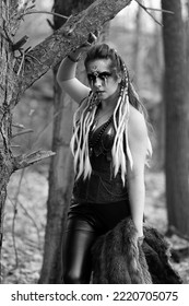 Black And White Portrait Of A Female Viking Wearing Face Paint, Fur, And Her Hair In Braids. She Is Standing In The Woods. 