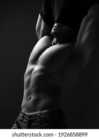 Black And White Portrait Of Handsome Muscular Man, Athlete With Perfect Built Body Standing Taking Off His Shirt, Getting Naked, Demonstrating Perfect Abs And Chest Muscles Over Dark Background