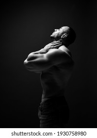 Black and white portrait handsome sexy muscular man, sportsman, athlete with perfect built body standing side to camera holding hand at neck, head thrown back and eyes closed over dark background