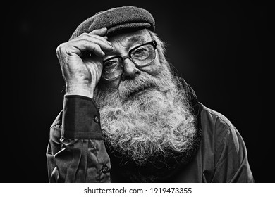 Black And White Portrait Of An Intelligent Tired Old Man In Glasses And Cap With A Long Gray Beard Looks At The Camera. Old Age Wisdom. 