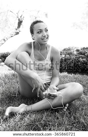 Black and white portrait of joyful african american teenager girl smiling using smartphone to listen to music, sitting on grass in park, outdoors. Adolescent technology lifestyle, exterior holiday.