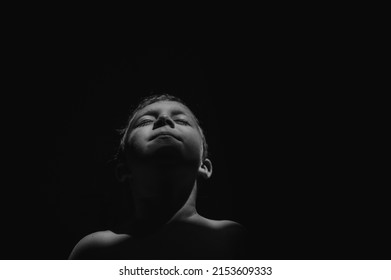 Black And White Portrait Of Little Boy With Eyes Closed Raising Face On Black Background