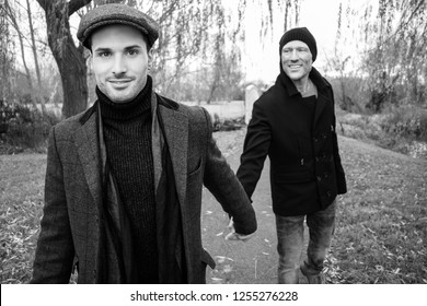 Black And White Portrait Of Male Gay Couple Holding Hands And Smiling As They Walk On Park Pathway