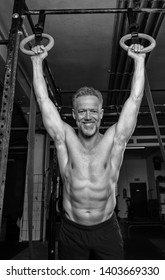 Black And White Portrait Of A Middle Aged Athlete Who Is Doing Strength Training In A Gym. The Sporty Man In His Fifties Is Shirtless And Hanging In The Rings. Fit Older Man With Gray Hair And Beard.