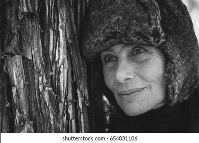 Black And White Portrait Of Middle Aged Woman Leaning On Tree Trunk Wearing Faux Fur Winter Hat With Flaps. Closeup On Mature Lady In Forest. Winter Fashion Sales Concept