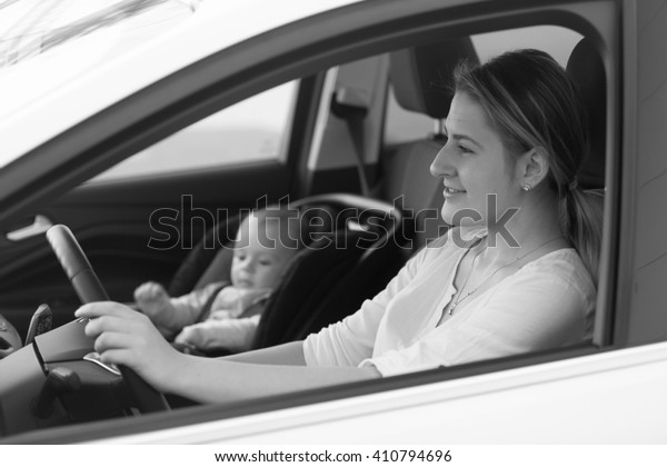 Black and white portrait of mother\
driving car with little baby boy sitting in safety\
seat