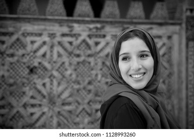 Black and white portrait of Muslim woman smiling in traditional clothing with hijab and black dress in front of traditional arabesque decorated wall