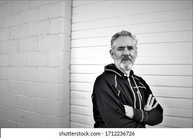 Black And White Portrait Photo Of A Fit, Attractive Older Caucasian Man, Looking At Camera. Short Beard And Gray Hair. Looks Calm And Determined.  Patterned Grey Wall Background.  Black Jacket.