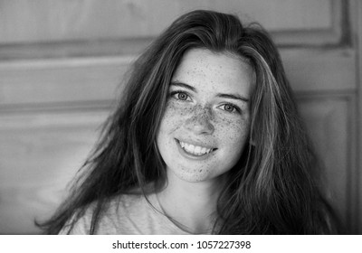 A Black And White Portrait Of A Positive And Happy Beautiful Girl With Freckles On Her Face, Looking With A Kind Smile At The Camera On The Background Of A Wooden Door. Positive And Happy Concept