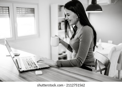 Black And White Portrait Of A Pretty Middle Aged Woman Working With Laptop While Drinking Cup Of Coffee In The Kitchen At Home. Home Office. In The Morning Concept.