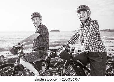 Black And White Portrait Of Senior Caucasian Couple Riding Off Road On The Pebble Beach With Bicycles At Sunset. Authentic Elderly Retired Life Concept