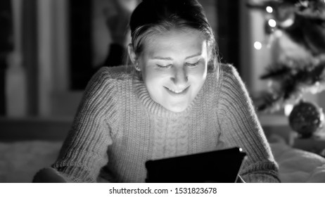 Black And White Portrait Of Smiling Woman With Tablet Lying On Floor At Night Under Glowing Christmas Tree