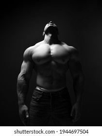 Black and white portrait of tall muscular man, athlete with perfect built body standing in jeans and shirtless with head thrown back, looking up over dark background