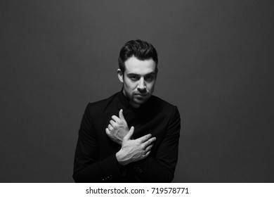 Black And White Portrait Of A Young Handsome Man In All Black Suit, Hands Crossed On The Chest, Looking At The Camera, Against Plain Studio Background.