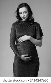 Black and white portrait of young pretty pregnant woman on gray studio background. Female in grey sequin dress with hands near pregnant belly.