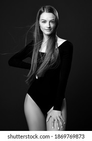 Black and white portrait of young slim woman with long silky straight fluttering in the wind hair in bodysuit holding hand on her waist and hip over dark background. Haircare, beauty, wellness concept