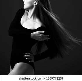Black and white profile of young beautiful woman with long silky straight flying fluttering hair in black bodysuit holding hand at her shoulder over dark background. Haircare, beauty, wellness concept