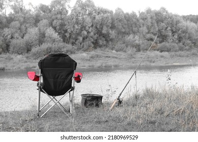 Black And White Retro Style Photo Of Red Fishing Chair Near Fishing Rod And Fishing Bait On River Bank In Summer On Fishing Trip