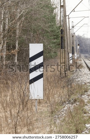 Black and white roadside marker post beside railway line in winter, reflective warning sign near tracks and overhead power poles.