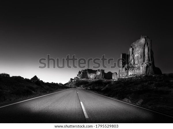 Black and white scene of a\
lonely road in Arches National Park Utah, at night. There is a\
large rock outcropping to the right and the sun is just visible to\
the left. 
