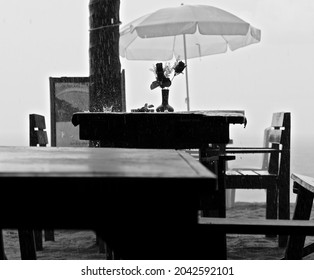 Black And White Scene On A Beach On A Rainy Day, A Table Under A White Umbrella With Two Empty Chairs Facing Each Other, In The Center A Bouquet Of Roses And A Drop Of Water Breaking. A Romantic Date.