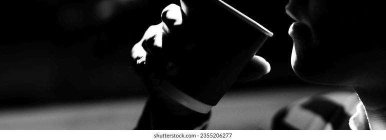 Black and white side view dramatic light shot of young woman holding paper cup with espresso on black background. Copy space