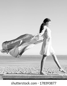 Black And White Side View Of An Elegant Woman Rising A Floating Silk Fabric Up With Her Arms Against A Sunny Sky, Traveling On A Holiday Beach. Taking A Step On A Stone Wall, Beauty And Lifestyle.