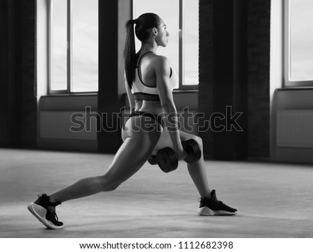 Black and white sideview of athletic girl performing fallouts keeping dumbbells in spacy gym with panoramic windows. Having strong, fit body with heatlthy tanned skin, muscles. Doing exercises.