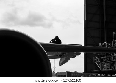 Black And White Silhouette Of Aircraft Technician Sitting On Upper Wing Of Airplane While Adjust And Maintenance Flight Control For Safety Before Return To Flight At Aircraft Hangar.